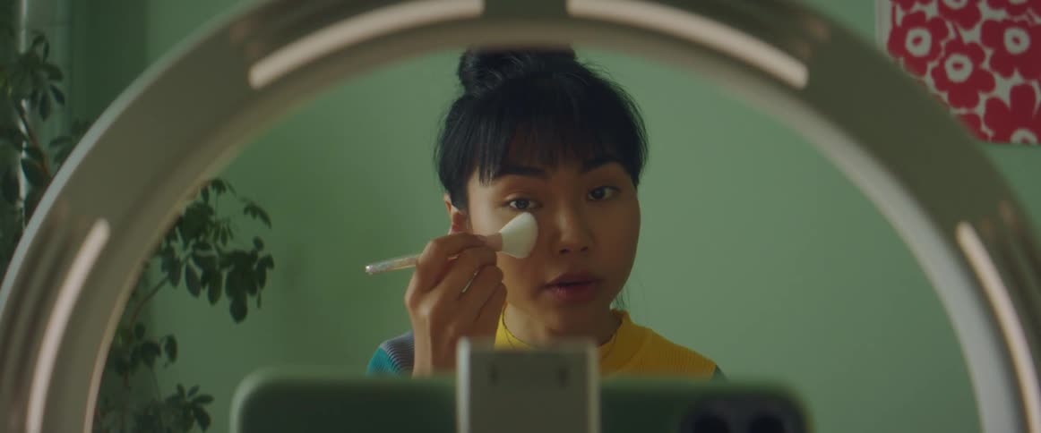 Woman with colorful shirt and earrings puts on makeup while filming herself in front of green wall