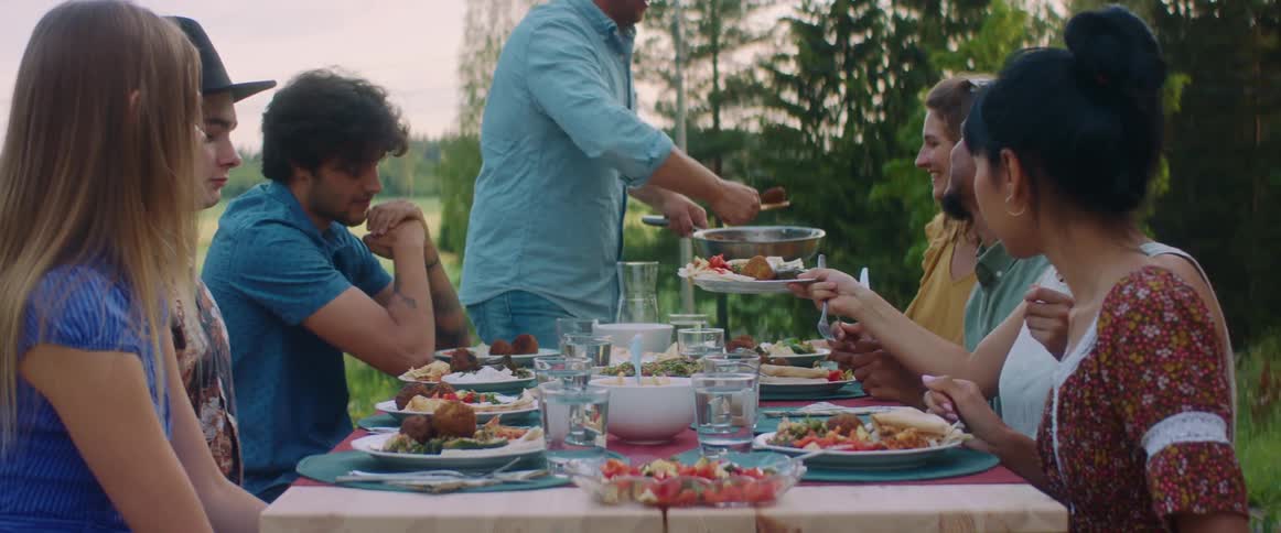 Man serves friends sitting at outdoor picnic table as they laugh and talk