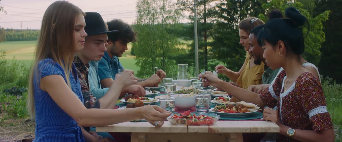 A group of friends serve themselves and each other food while sitting at a picnic table in the yard
