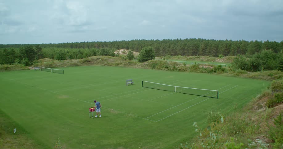 A man walks toward a tennis court on land surrounded by a forest