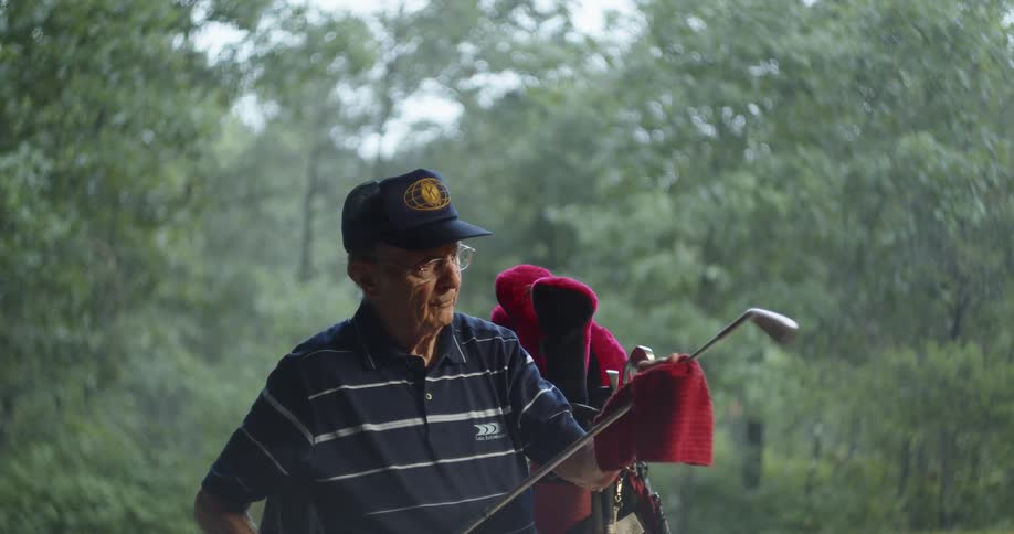Man wipes golf club with cloth standing next to full golf bag with trees in background