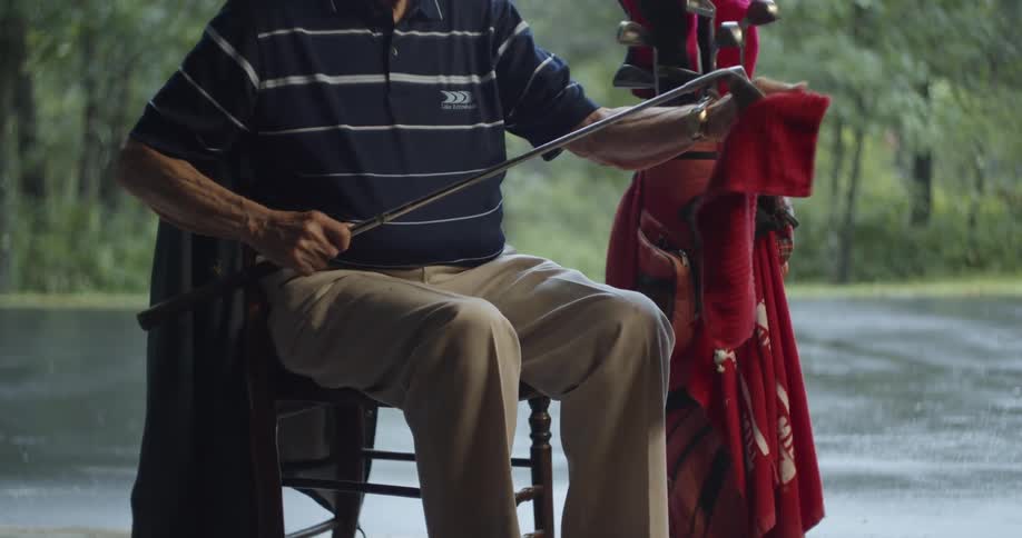 Elderly man sits in a chair outdoors and washes a golf club with water and a red cloth