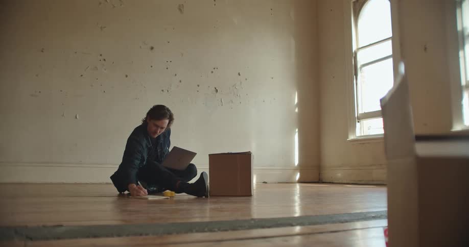 A man in a room sitting on a floor writing with a pen and using a laptop with a wall in the background