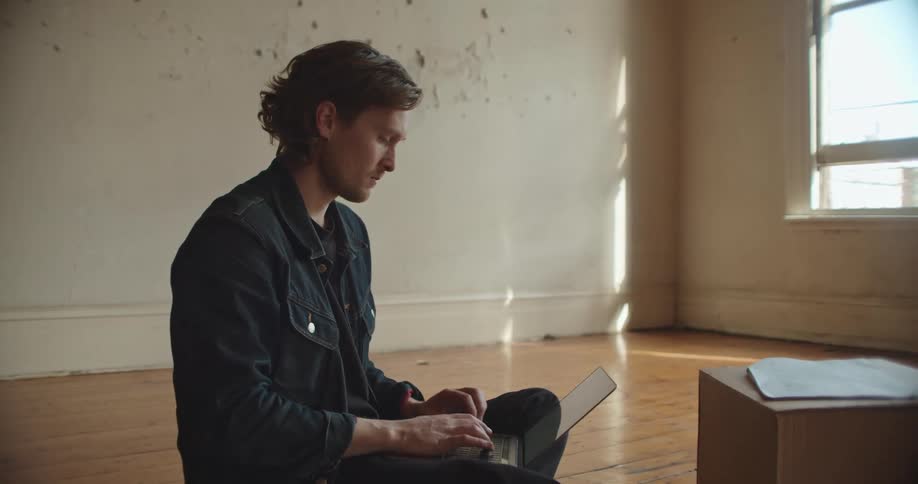 Man with denim jacket sitting on floor types on laptop in empty room