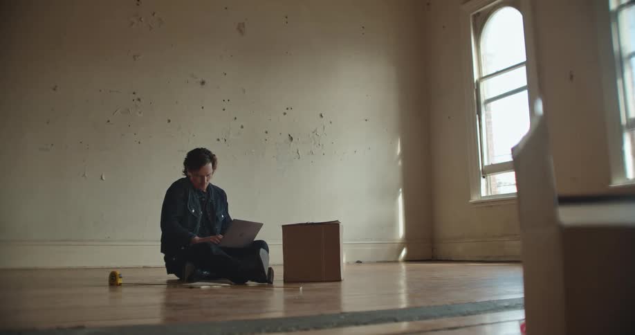 A man sitting on the floor of an empty room types on the laptop resting on his laptop while holding a smartphone to his face with his shoulder
