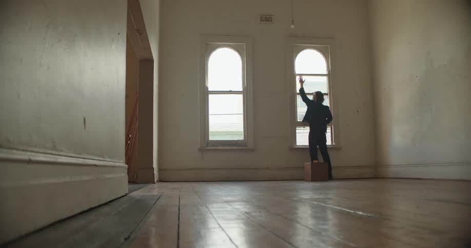 Man with denim jacket and pants carries a box and opens window in empty room