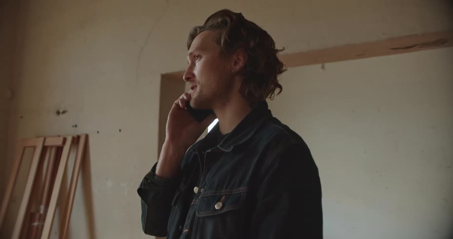 A man in a room talking on a smart phone with a doorway a window and a wall in the background