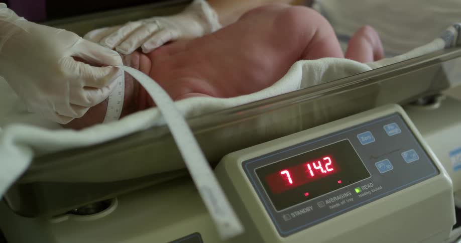 In a hospital a gloved nurse is measuring the head of an infant and weighing its body