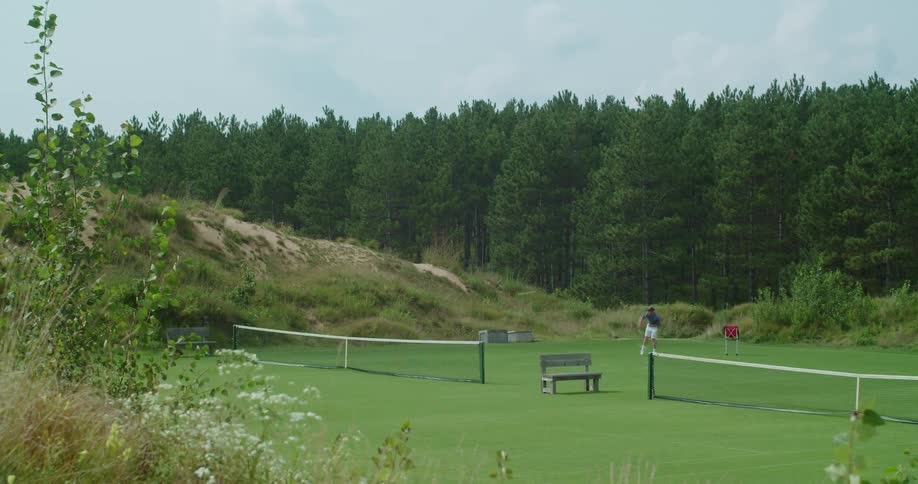 A man plays tennis alone in a court next to a forest