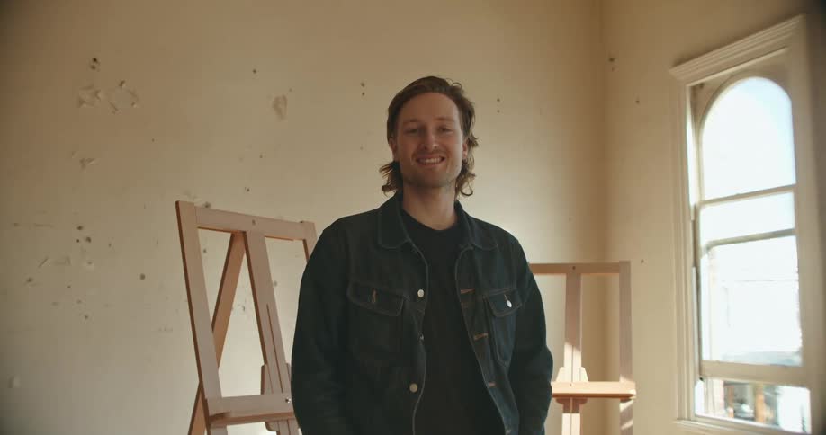 A man stands in a room with empty easels and puts his hands in his pockets and smiles