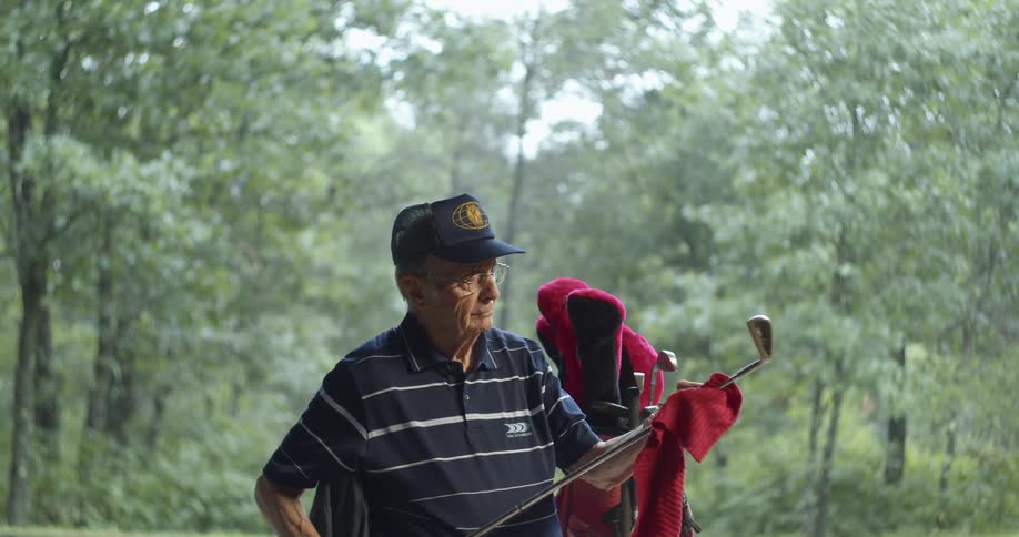 Older man stands near full golf bag cleaning a golf club with a rag with woods as a backdrop