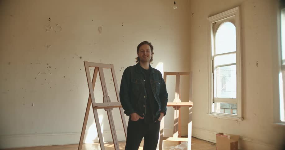 Artist stands smiling in empty apartment near two empty easels
