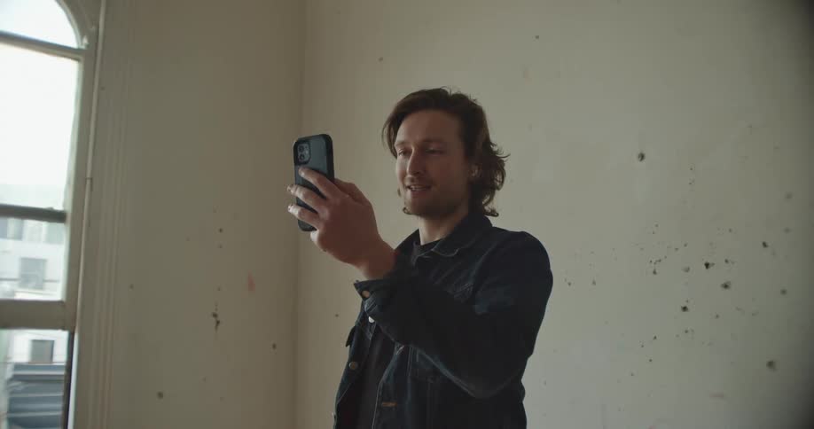A man standing near a window in an apartment waves at his phone on a video call as he talks