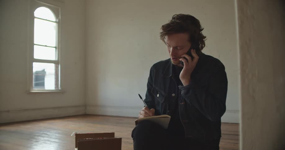 A man is conducting business on a smartphone and taking notes while kneeling in an empty apartment