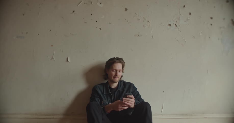 A man smiling while scrolling on a smartphone and leaning against a wall
