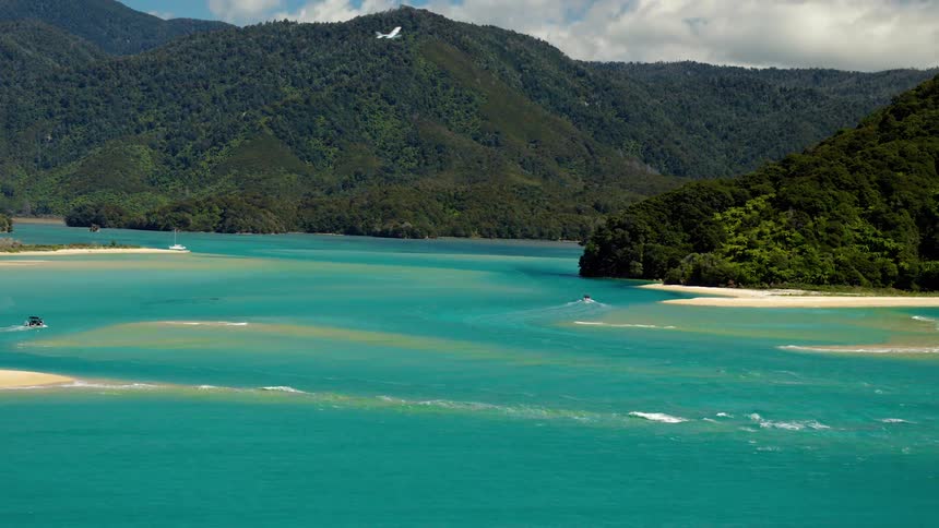 Boats cut through azure blue water as airplane flies overhead near forest covered mountains