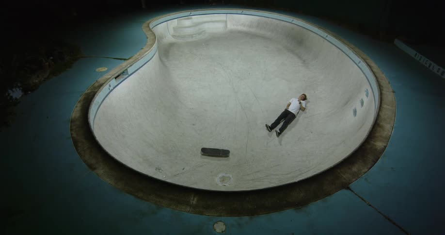 A man lying alone in an empty swimming pool with a skateboard at night