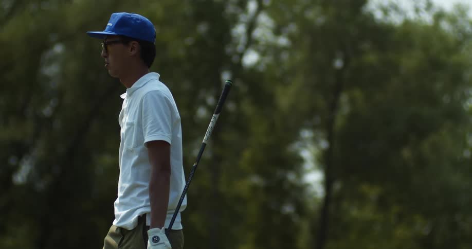 Man with blue cap glasses white shirt and shorts carries golf club walking around