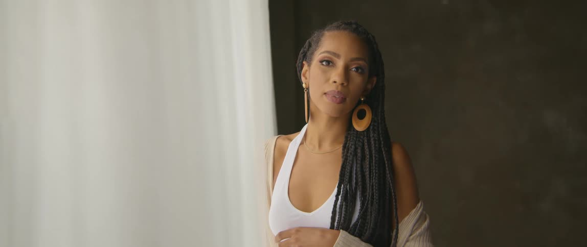 A woman with long braids and large wooden earrings turns her head as she poses