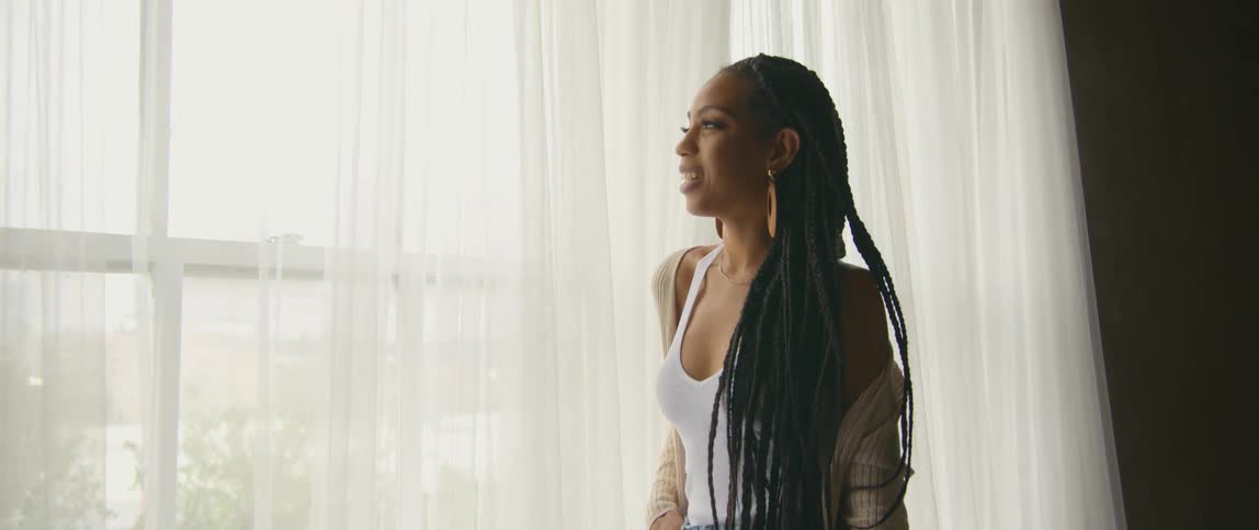 Woman with earrings and braids looks out the window with curtains