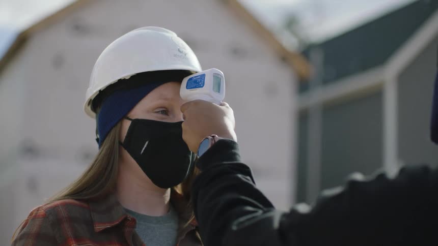 A woman with a hardhat and a mask getting her temperature check with a thermometer by another person with houses the sky and clouds in the background