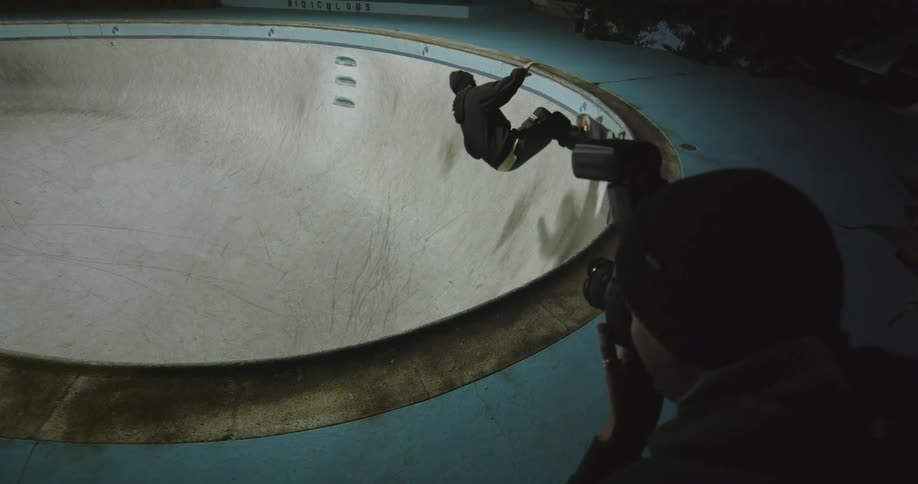 Film photographer takes picture of skateboarder riding skateboard along bottom and sides of empty swimming pool at night