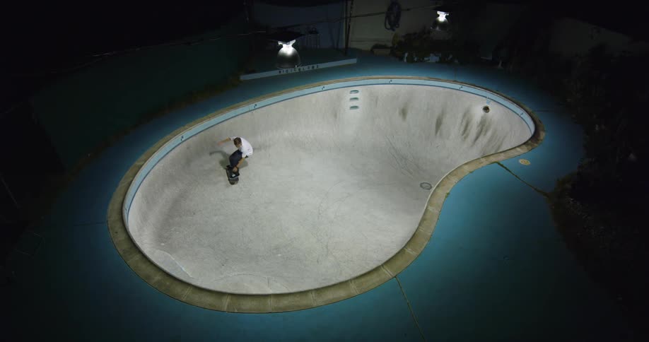 Skateboarder skates around bottom and sides of empty swimming pool in dark are lit by overhead light
