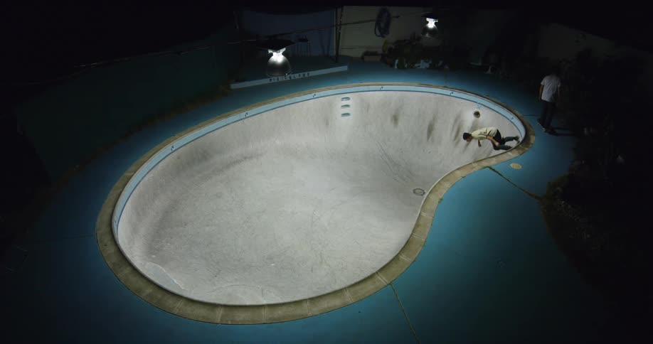 A skateboarder rides in an empty pool while another skateboards around the pool deck