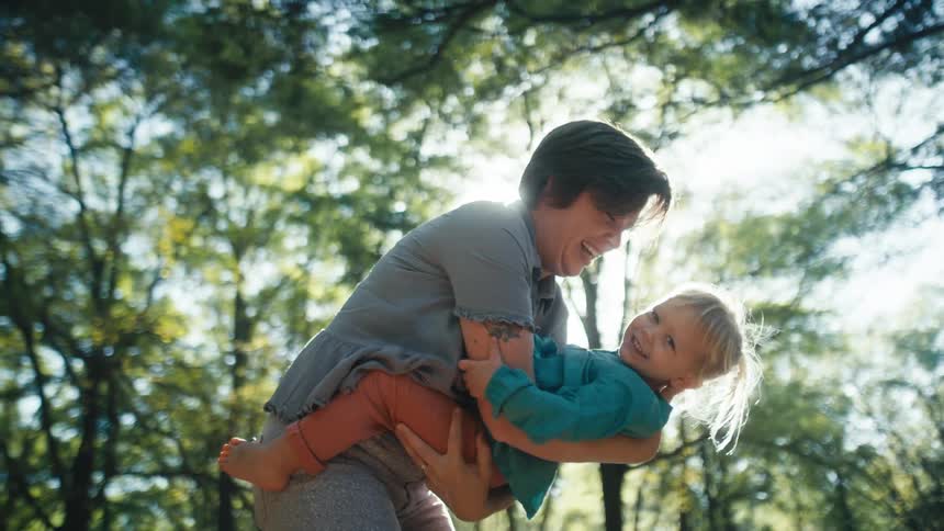 A woman holds her daughter and tilts her forward as they play in a forest