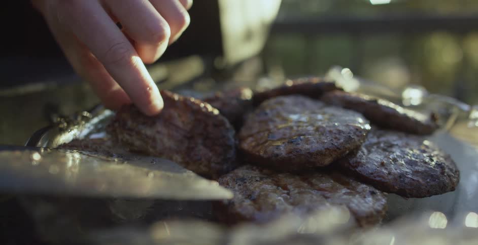 A person picks up freshly coocked burgers from a plate near a barbecue at evening