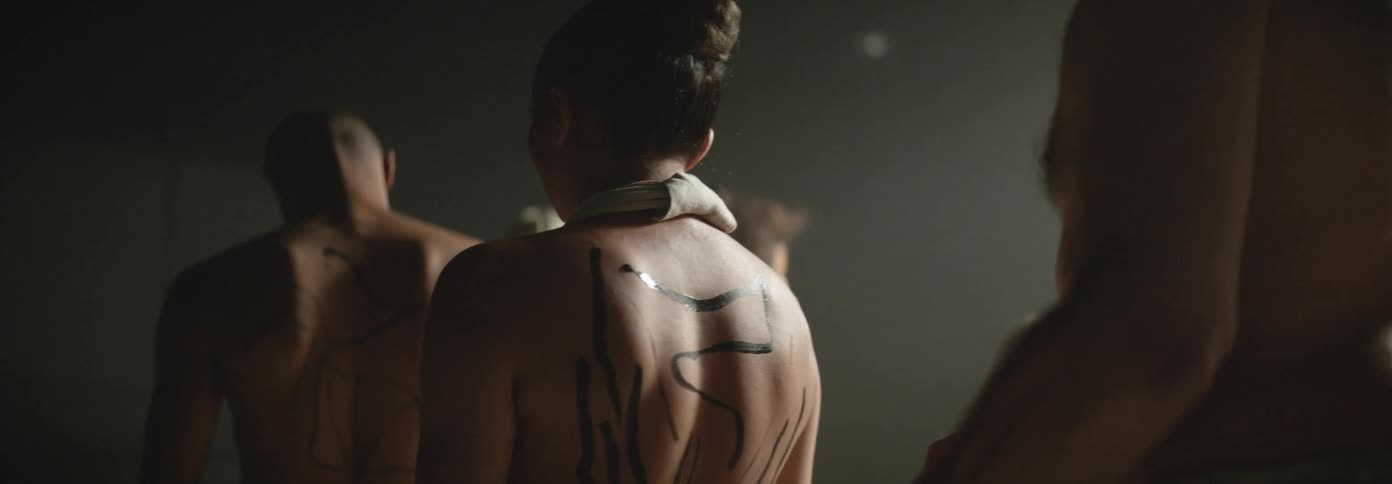 Group of naked people with ink tattoos on their backs standing in a circle inside a dimly lit room
