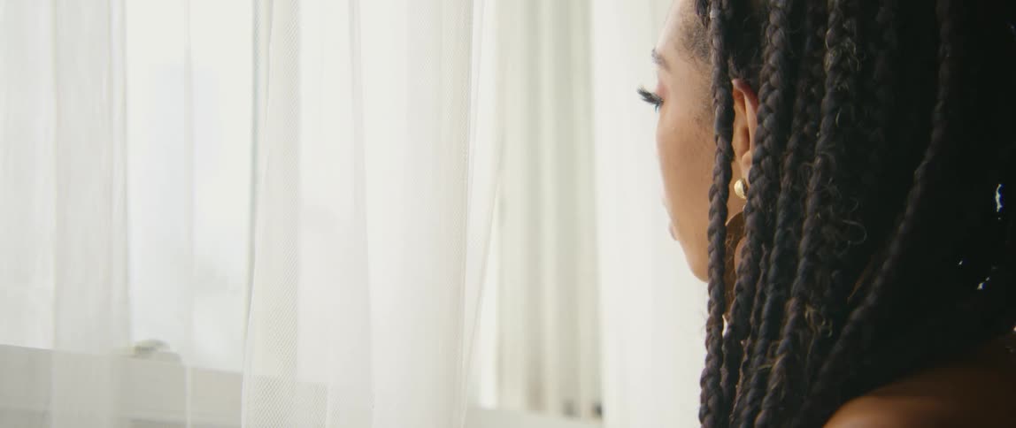 A woman in a room standing next to a window with curtains with a wall in the background