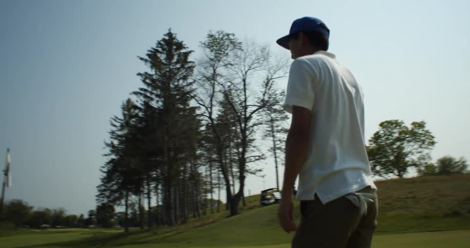 A man with a golf club walking on a golf course with trees the sky and clouds in the background