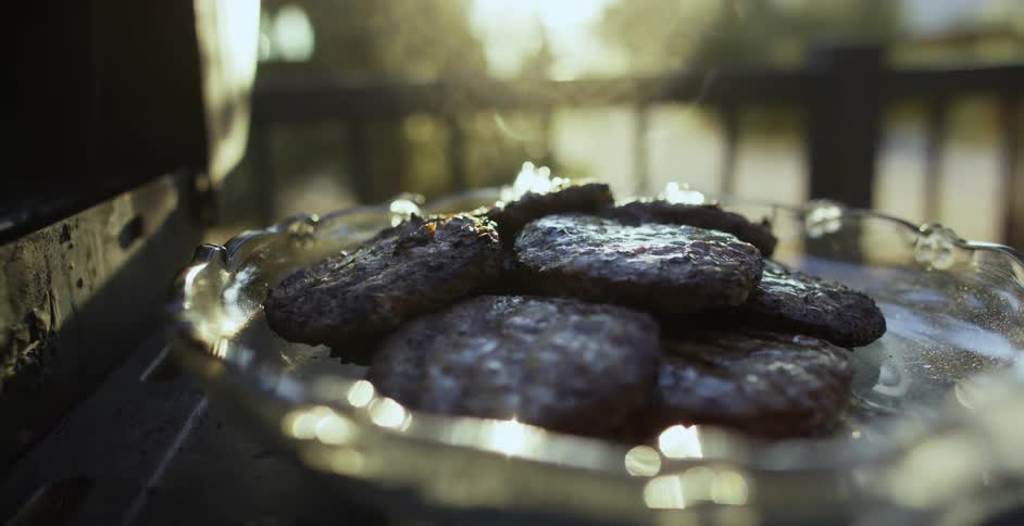 Smoke rises from a plate of burger patties next to a grill outdoors