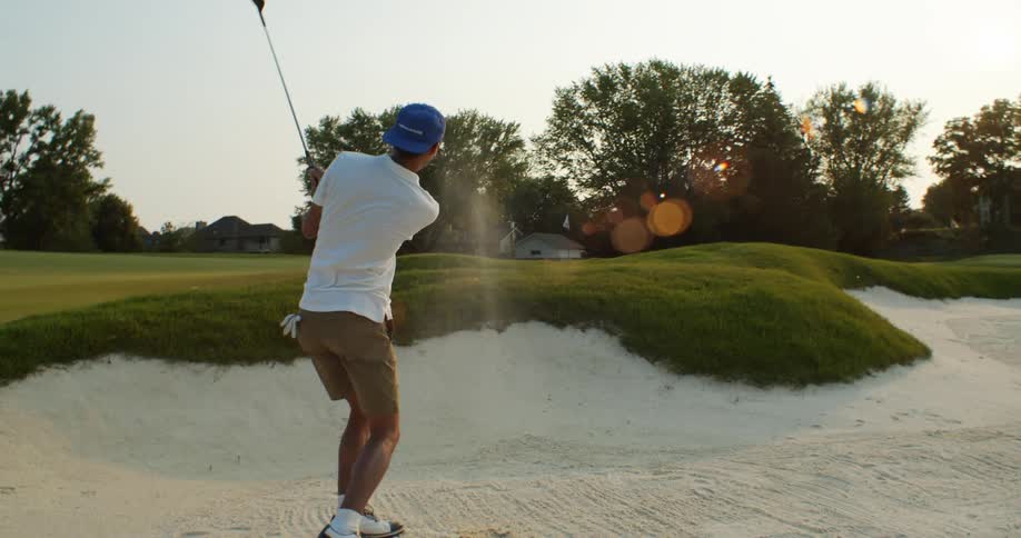Golfer takes stroke hitting golf ball out of sand trap under clear sky