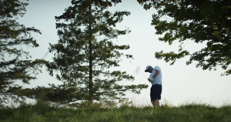 A golfer swings his club and hits the ball between trees and over a slope