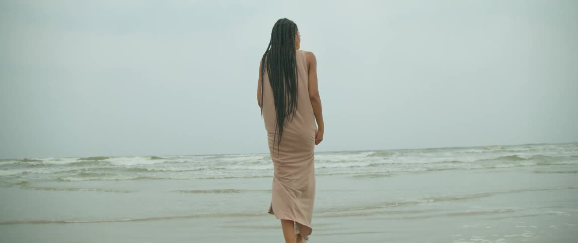 A woman wearing a long dress is walking along a beach on an overcast day with foaming waves in the background