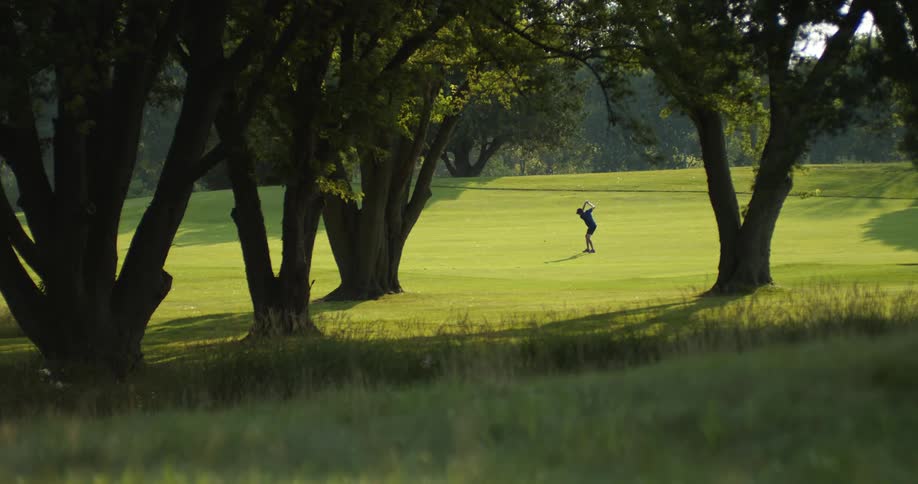 A man in a golf course playing golf with trees in the foreground and trees and the sky and clouds in the background