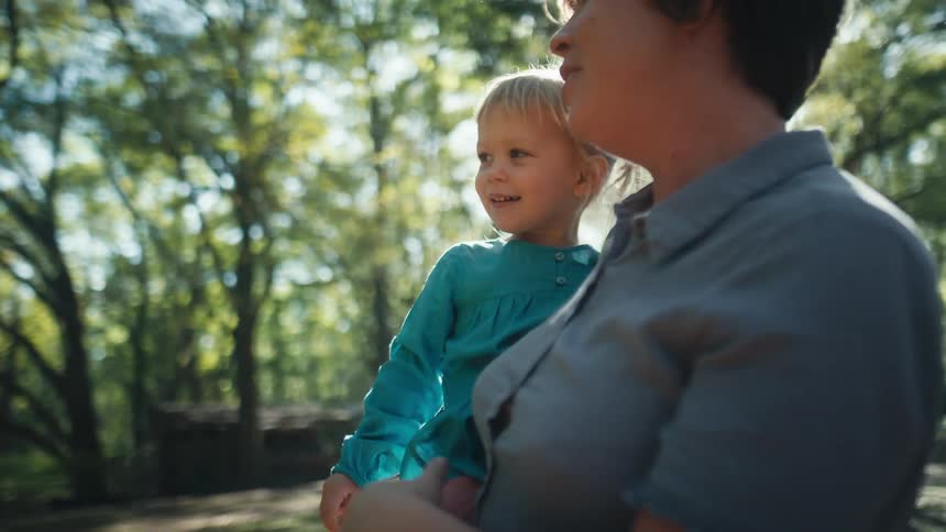 Mother holding daughter on her arms standing near the trees at morning