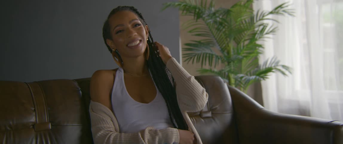Woman sits on sofa smiling as she strokes long hair braids