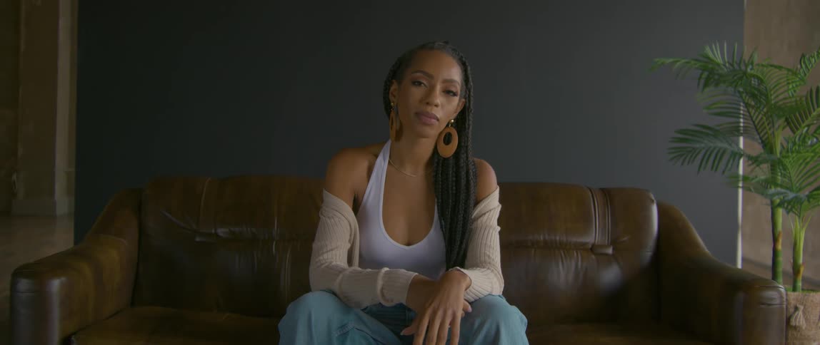 Woman with braids hoop earrings white tank top and jeans sits on brown sofa in front of dark wall and faces the camera with eye contact