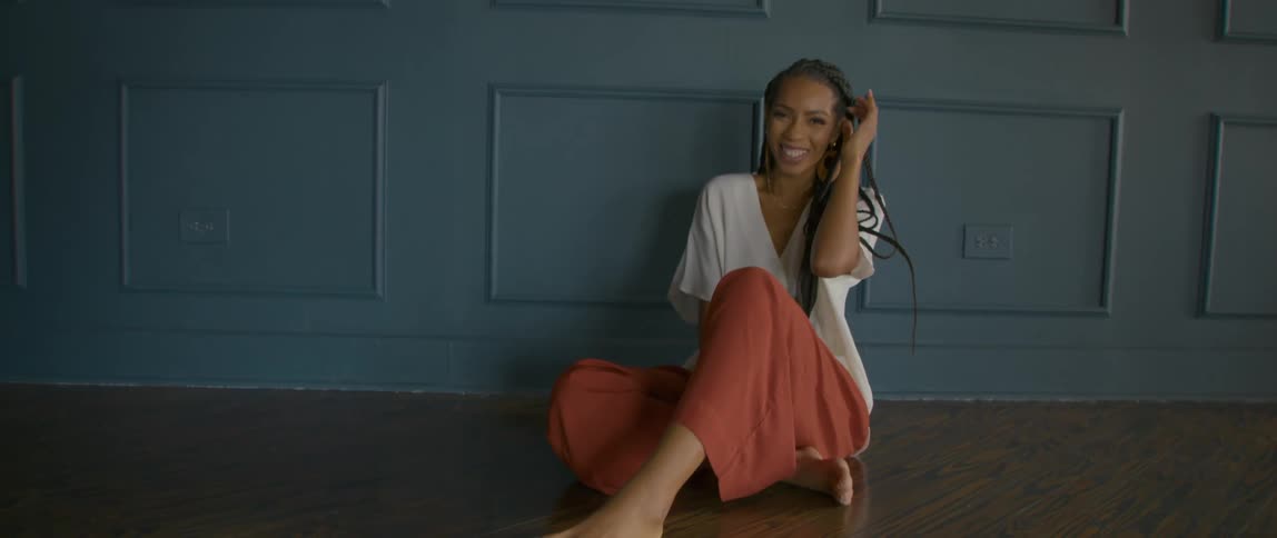 Woman with braids white blouse and pants sitting on floor smiles at camera in front of blue wall