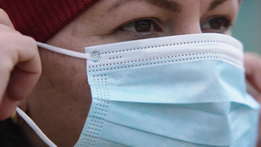 Woman wearing a beanie puts on a disposable blue face mask
