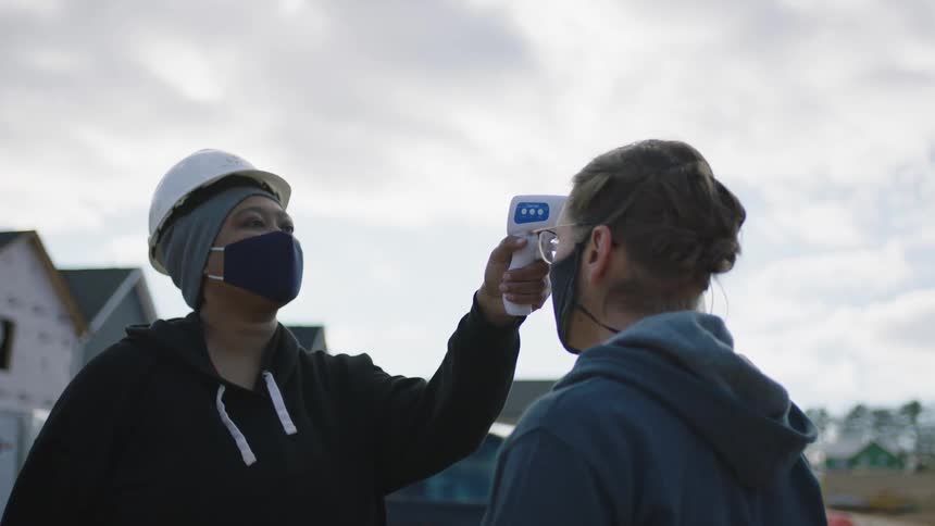 A woman on a home construction site uses a digital thermometer to measure another womans temperature before beckoning the woman to follow