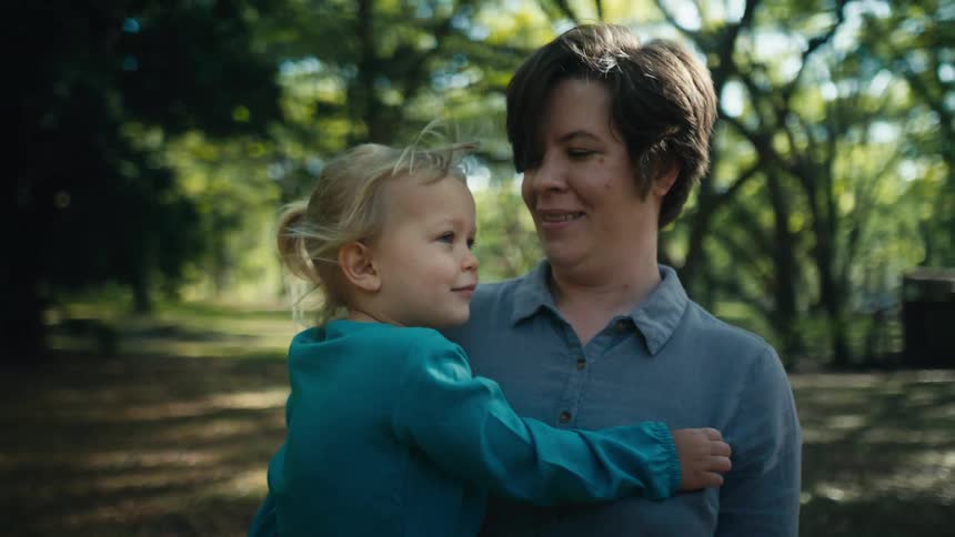 Woman holds blonde girl swaying as she stands in field surrounded by trees