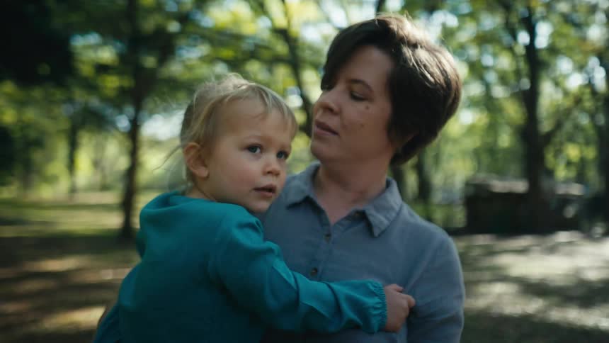 A small child is in arms of a lady who as they are standing outside surrounded by trees