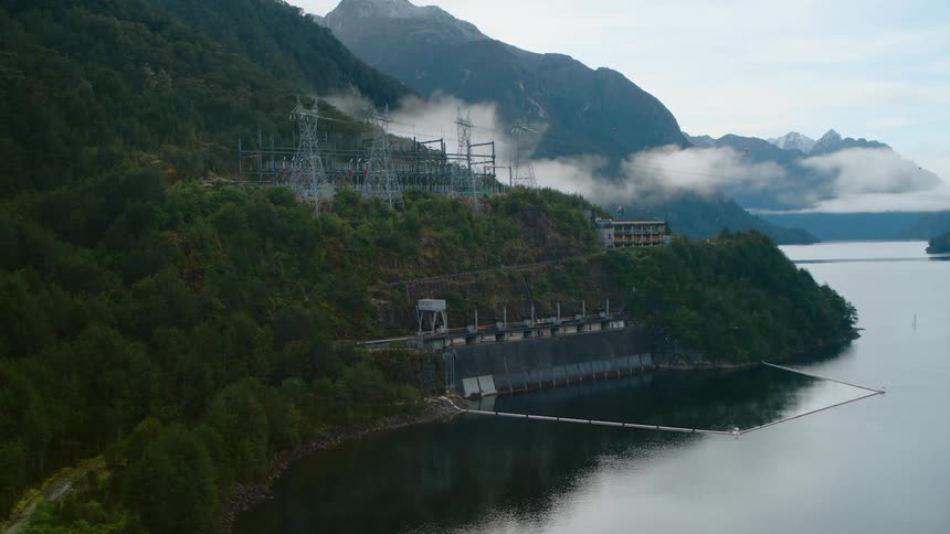 An aerial view of a power structure by a large lake with mountains and forest around it