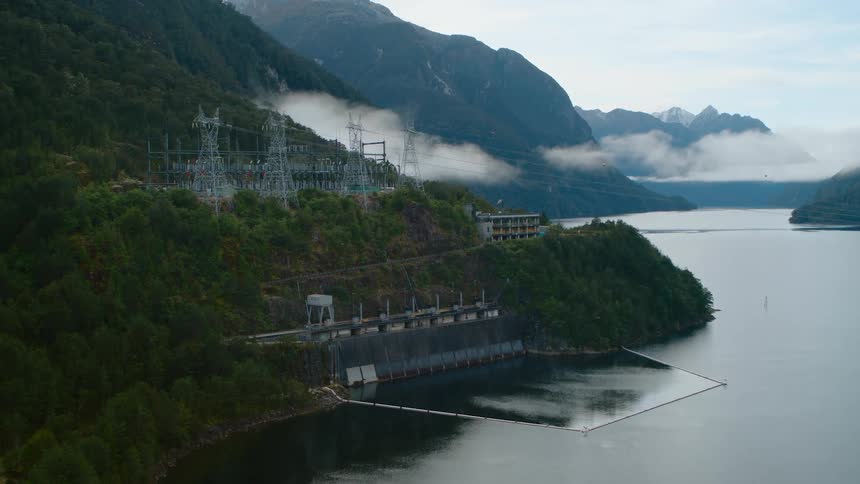 Low clouds float over power station sitting at foot of forest covered mountain near water