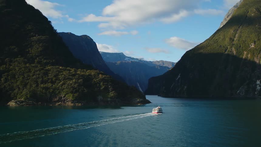 A boat is sailing on a lake between mountains and there are a few clouds in the sky