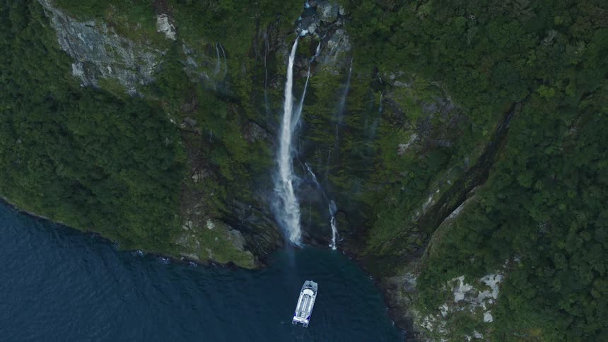 A large boat floats in the ocean in front of a waterfall descending a side of mountain on the coast